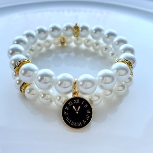 May include: A double-strand pearl bracelet with a gold clock charm. The bracelet is made of white faux pearls and has gold accents. The clock charm has Roman numerals and black hands.