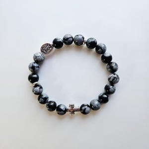 以下が含まれることがあります： 黒とグレーの斑点模様の石ビーズを使用したブレスレット。シルバーカラーのクロスチャームと装飾用のシルバーカラーのアクセントビーズが付いています。手首に巻いて着用するようにデザインされています。