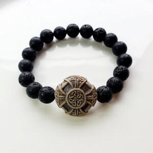 May include: Black lava stone bead bracelet with a decorative, weathered-look, Celtic cross centerpiece. The bracelet features round, porous black beads and a detailed, tan-colored cross with intricate knotwork.