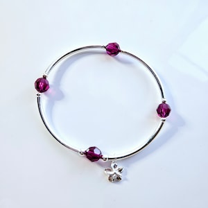 May include: A silver bangle bracelet featuring five deep magenta crystal beads and a small, silver flower charm. The bracelet has a simple, elegant design. The beads are evenly spaced around the bracelet, suitable for everyday wear.