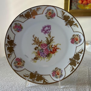 May include: White porcelain plate with a gold rim and floral design. The plate features a central bouquet of pink, purple, and red flowers, surrounded by smaller floral clusters and gold leaf accents. A decorative piece for serving.