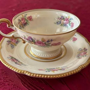 Può includere: Servizio da tè color crema con disegni floreali rosa, viola e blu. La tazza e il piattino hanno bordi dorati e un bordo smerlato. Anche il manico della tazza è dorato.