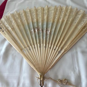 May include: A folding hand fan with cream-colored ribs and a lace-trimmed upper section. The center features a painted scene of cherubs against a pale blue background. A small metal ring with a decorative tassel is attached to the base of the fan.