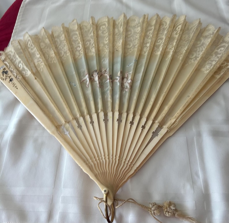 May include: A folding hand fan with cream-colored ribs and a lace-trimmed upper section. The center features a painted scene of cherubs against a pale blue background. A small metal ring with a decorative tassel is attached to the base of the fan.