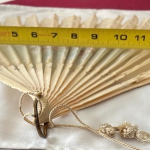 May include: A cream-colored folding hand fan with a delicate, pleated design and a small tassel attached to the base. A yellow measuring tape is positioned across the top, indicating a width of approximately 15 inches when fully extended.