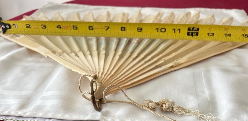 May include: A cream-colored folding hand fan with a delicate, pleated design and a small tassel attached to the base. A yellow measuring tape is positioned across the top, indicating a width of approximately 15 inches when fully extended.