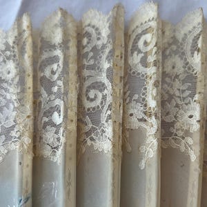 May include: A folding hand fan featuring cream-colored lace with intricate floral and scrollwork patterns. The lace is attached to vertical, light-colored slats that form the structure of the fan, creating a pleated, layered appearance throughout.