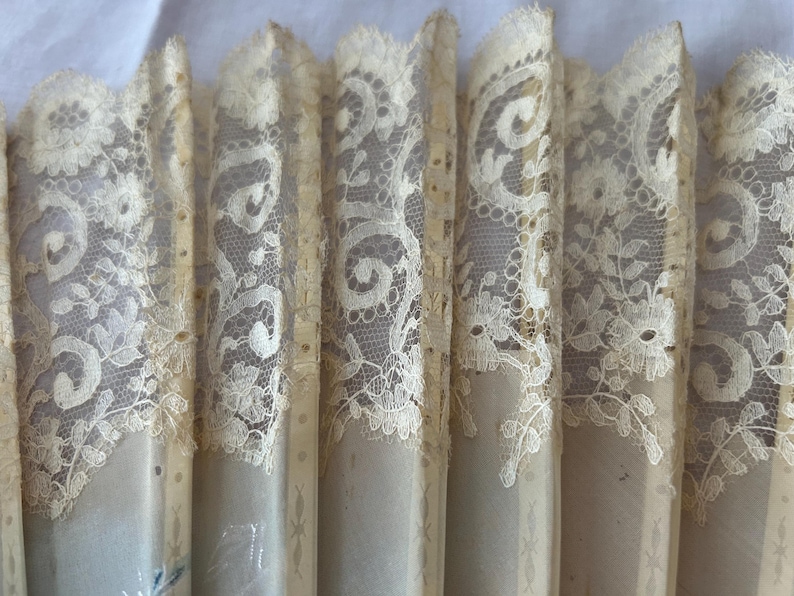May include: A folding hand fan featuring cream-colored lace with intricate floral and scrollwork patterns. The lace is attached to vertical, light-colored slats that form the structure of the fan, creating a pleated, layered appearance throughout.