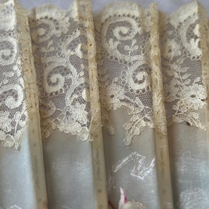 May include: A folding hand fan featuring cream-colored fabric panels overlaid with intricate floral lace trim. The fan structure consists of vertical slats with subtle decorative lines, showcasing a delicate texture and a soft, neutral color palette.