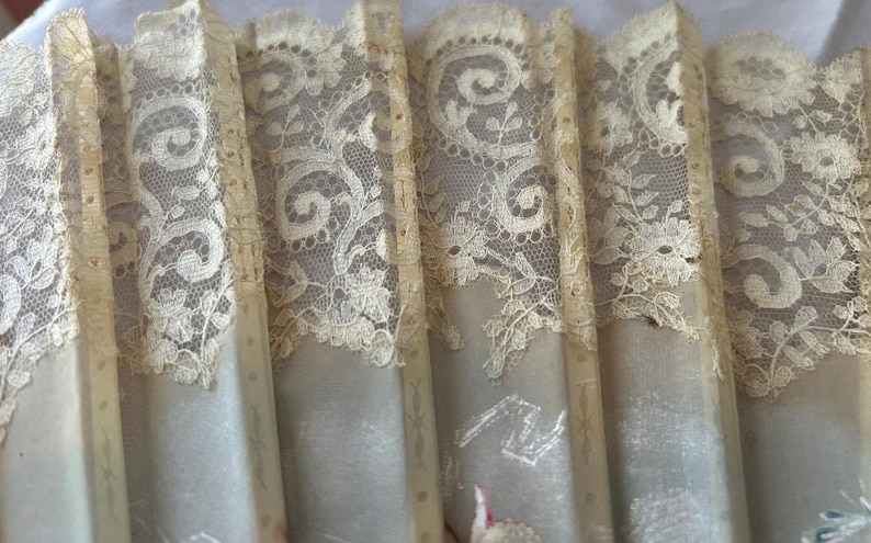 May include: A folding hand fan featuring cream-colored fabric panels overlaid with intricate floral lace trim. The fan structure consists of vertical slats with subtle decorative lines, showcasing a delicate texture and a soft, neutral color palette.