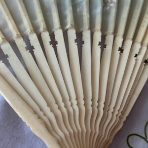 May include: A folding hand fan featuring off-white, carved, rigid slats that radiate from a central pivot point. The upper portion of the fan consists of a translucent, pale fabric leaf attached to the top of each individual carved slat.