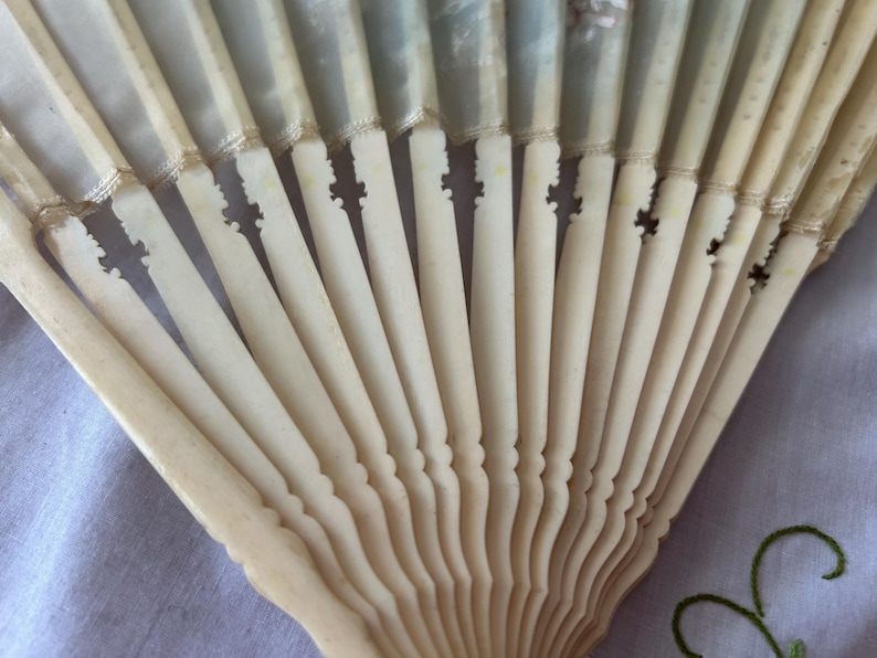 May include: A folding hand fan featuring off-white, carved, rigid slats that radiate from a central pivot point. The upper portion of the fan consists of a translucent, pale fabric leaf attached to the top of each individual carved slat.