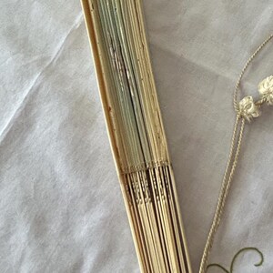 May include: A folded hand fan constructed from light-colored, thin wooden slats. The fan is secured at the base with a circular metal ring and features a decorative off-white cord with small knotted tassels attached to the handle area.