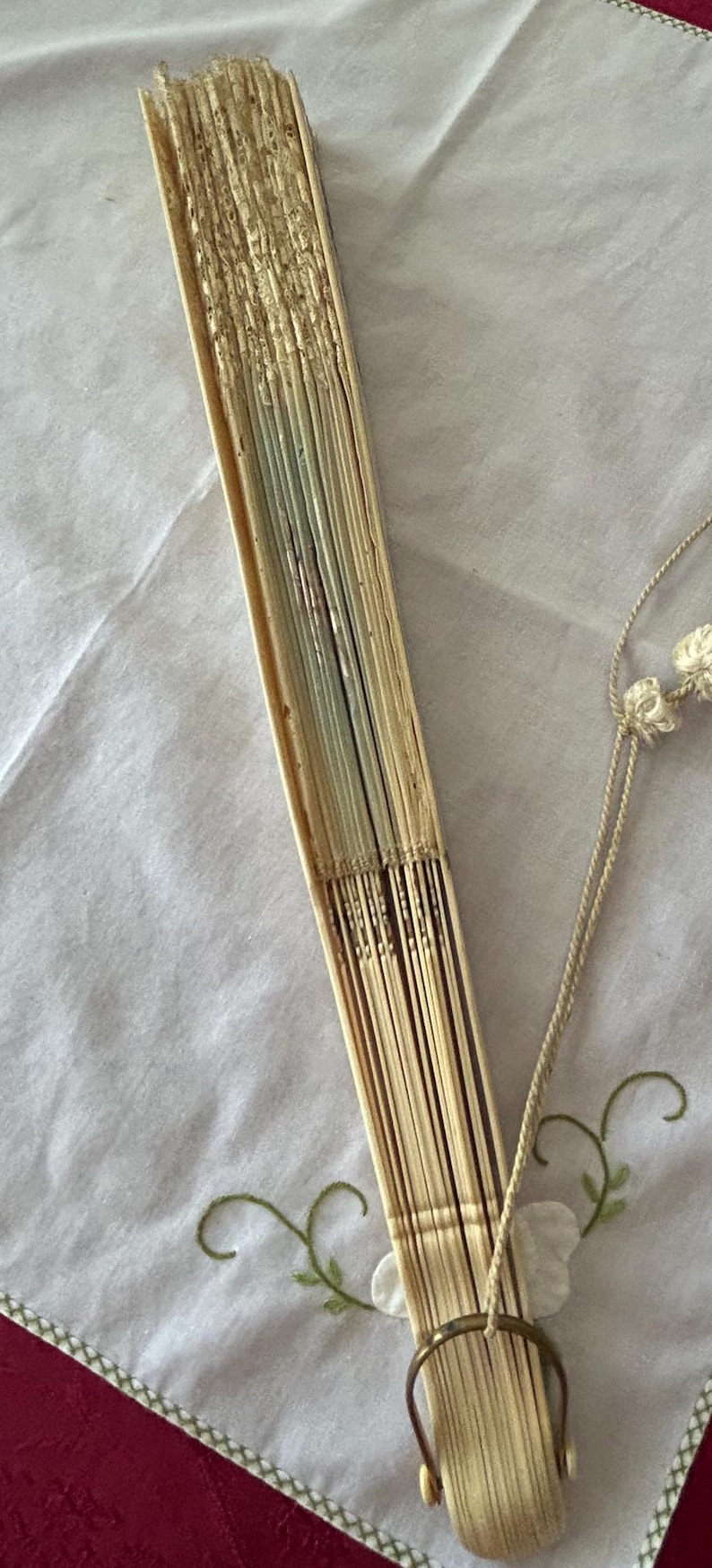 May include: A folded hand fan constructed from light-colored, thin wooden slats. The fan is secured at the base with a circular metal ring and features a decorative off-white cord with small knotted tassels attached to the handle area.