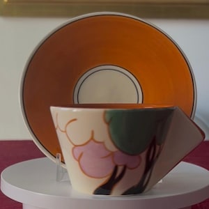 May include: A decorative teacup and saucer set. The saucer is orange with a white and black inner circle. The teacup is cream-colored with a geometric design featuring pink and green floral elements. The cup has a unique angular handle.