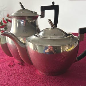 May include: Two silver teapots with black handles and lids. The teapots are on a red tablecloth. One teapot is taller than the other. The handles are a dark, rectangular shape. The spouts are curved.