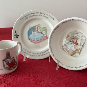 May include: A white ceramic mug, plate, and bowl set featuring illustrations of Peter Rabbit. The mug and plate have text. The set is displayed on a red tablecloth.