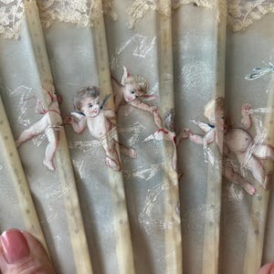 May include: A folding hand fan featuring a light gray fabric surface decorated with painted cherubs and small insects. The fan has vertical ribs and a delicate cream-colored lace trim along the top edge, creating a detailed and ornate appearance.