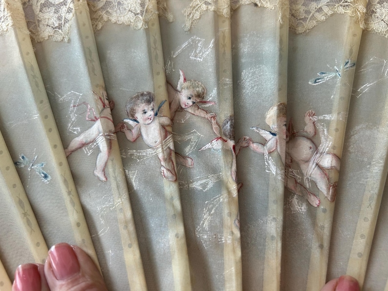 May include: A folding hand fan featuring a light gray fabric surface decorated with painted cherubs and small insects. The fan has vertical ribs and a delicate cream-colored lace trim along the top edge, creating a detailed and ornate appearance.