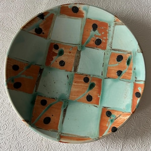 May include: A handmade ceramic plate with a unique design. The plate features a checkerboard pattern of orange squares and light blue squares. Each orange square has a black dot and green paint streaks. The plate is round.