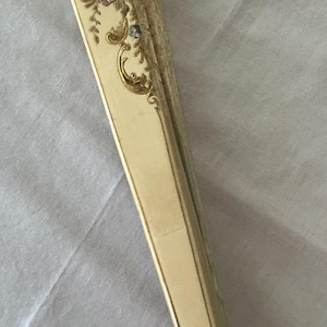 May include: A folded hand fan with a cream-colored handle and frame. The top section features intricate floral patterns in shades of pink, blue, and green, accented with gold-colored scrollwork details. The base of the handle displays a small floral motif.