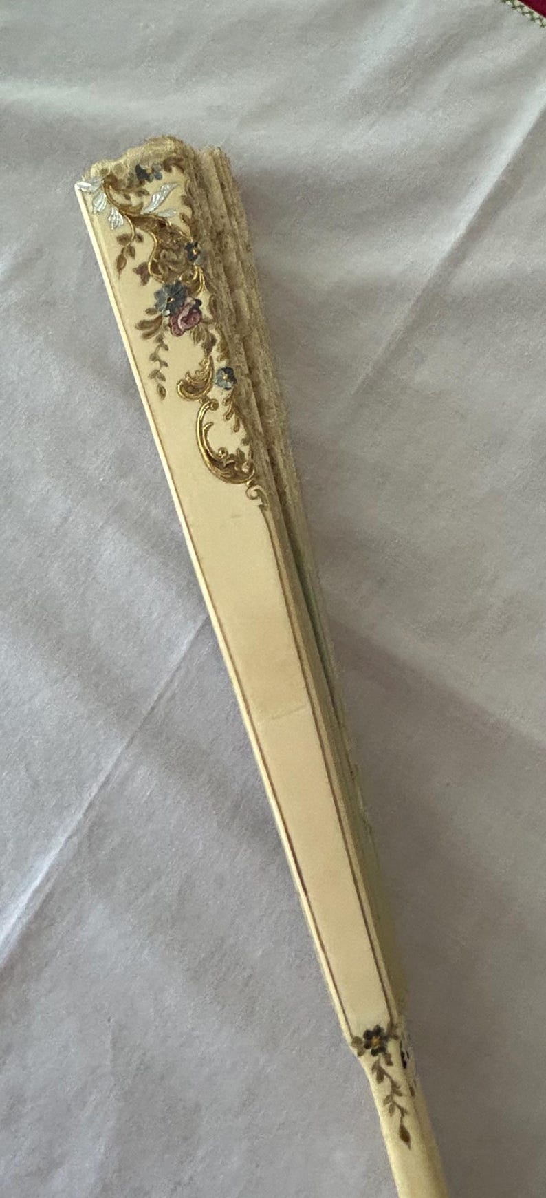 May include: A folded hand fan with a cream-colored handle and frame. The top section features intricate floral patterns in shades of pink, blue, and green, accented with gold-colored scrollwork details. The base of the handle displays a small floral motif.