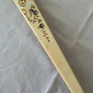 May include: A folded hand fan with a cream-colored handle featuring a decorative floral motif. The design includes small dark blue flowers with gold stems and leaves, accented by a silver-toned scrollwork pattern near the top edge of the fan.