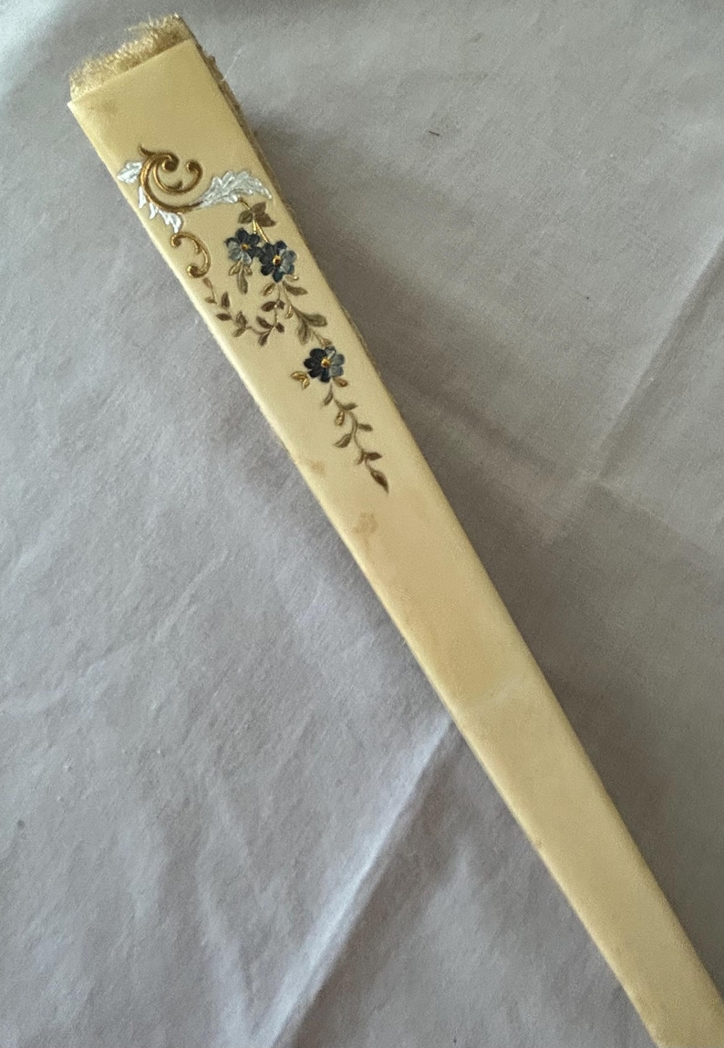 May include: A folded hand fan with a cream-colored handle featuring a decorative floral motif. The design includes small dark blue flowers with gold stems and leaves, accented by a silver-toned scrollwork pattern near the top edge of the fan.