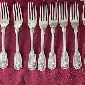 May include: A set of eight silver-toned metal forks arranged in a row. Each fork features four tines and a decorative handle base with a raised shell motif. The handles have a vertical ribbed design extending from the neck to the base.