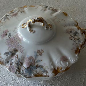 May include: White porcelain serving dish with lid, featuring a floral design in pink, blue, and gray. The dish has gold trim and handles, and a decorative handle on the lid. A vintage piece.