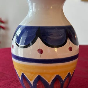 Puede incluir: Un jarrón de cerámica con la parte superior blanca y un diseño colorido. El jarrón presenta bandas azules, amarillas y rojas, con patrones decorativos. El jarrón mide aproximadamente 15 cm de alto, con un cuerpo redondeado y una abertura acampanada.