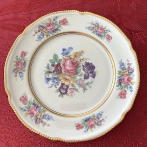 May include: A white porcelain plate with a gold rim and a floral design. The plate features a central bouquet of pink, purple, and yellow flowers, with smaller floral clusters around the edge. The plate has a scalloped edge.