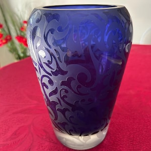 Puede incluir: Un jarrón de vidrio azul cobalto con un patrón grabado en espiral. El jarrón tiene un cuerpo ancho y redondeado que se estrecha hacia la base. El diseño grabado es de un color más claro que el jarrón en sí. El jarrón está sobre una superficie roja.