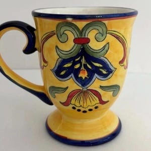 May include: Yellow ceramic mug with a pedestal base and a curved handle. The mug is decorated with a floral design in blue, green, and red. The rim and handle are trimmed in blue.