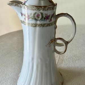 Puede incluir: Cafetera de porcelana blanca con tapa, con un diseño floral en rosa, verde y azul. La cafetera tiene un borde y un asa dorados, y un cuerpo acanalado. Una pieza decorativa para servir café o té.
