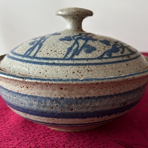 May include: A ceramic lidded bowl with a speckled, light gray finish. The bowl features blue horizontal stripes and floral designs on the lid. The lid has a small knob for easy handling. The bowl is likely used for serving or storage.