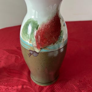 May include: A ceramic vase with a flared rim, featuring a white upper section and a brown lower section. The surface is decorated with a painted red floral motif, green leaves, and a horizontal teal band that separates the two main color zones.