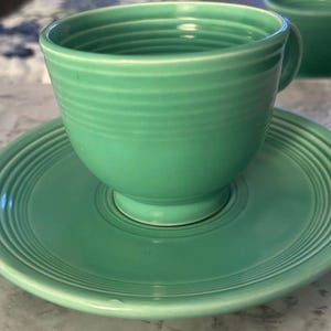 Puede incluir: Un juego de taza de té y platillo de cerámica en color verde claro. La taza presenta una forma redondeada con detalles de anillos concéntricos horizontales en el exterior. El platillo también muestra patrones de anillos concéntricos.