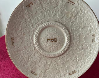May include: A white ceramic Passover Seder plate with a gold rim and embossed floral and scrollwork design. The plate features Hebrew text in gold and a central circular area with a crown and more Hebrew text. The plate is displayed on a clear stand.