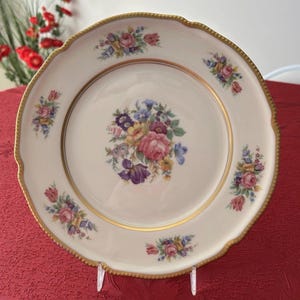 Può includere: Un piatto decorativo color crema con bordo smerlato e finiture dorate. Il piatto presenta una composizione floreale centrale e piccoli gruppi floreali attorno al bordo. I fiori sono nelle tonalità del rosa, viola, blu e giallo.