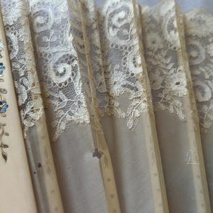 May include: A folding hand fan featuring cream-colored ribs and delicate beige lace panels. The leftmost rib is decorated with a painted floral motif in blue, gold, and silver tones, while the lace displays an intricate, repeating scrollwork pattern.
