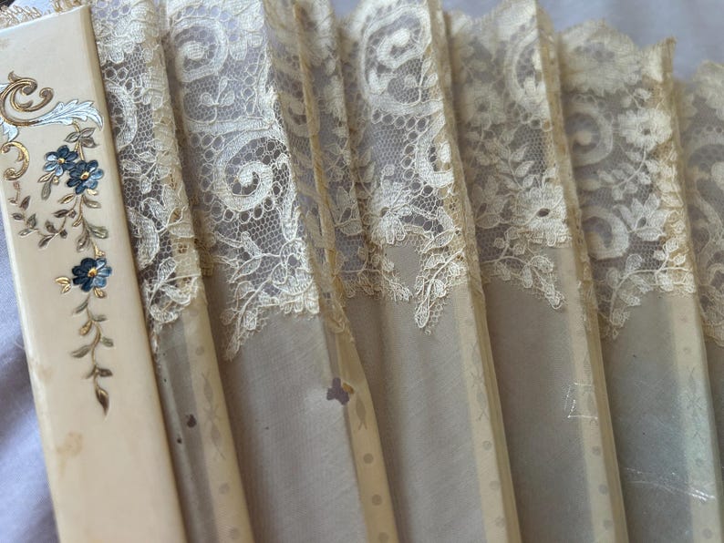 May include: A folding hand fan featuring cream-colored ribs and delicate beige lace panels. The leftmost rib is decorated with a painted floral motif in blue, gold, and silver tones, while the lace displays an intricate, repeating scrollwork pattern.