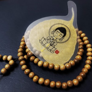 May include: A wooden prayer bead mala necklace with a clear leaf-shaped card featuring a cartoon image of a meditating figure. The beads are light brown and arranged in a circular pattern.