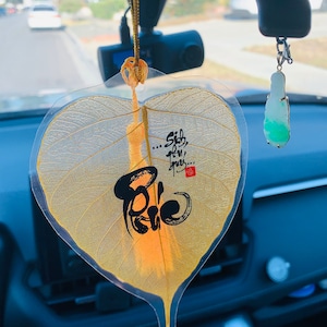 May include: A car interior shot features a leaf-shaped car charm with gold accents and a black script. The charm hangs from a gold chain. A jade pendant hangs from the rearview mirror.
