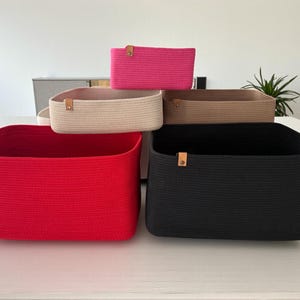 May include: A collection of rectangular storage baskets made from coiled rope in red, black, beige, tan, and hot pink. Each basket features a small, light brown leather-like tab attached to the side with a metallic rivet for a secure finish.