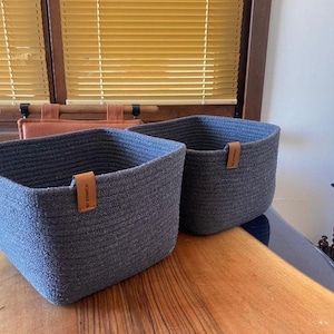May include: Two square storage baskets made of dark gray coiled rope with rounded corners. Each basket features a small, light brown leather-like tag attached to the upper rim, with the text "BY EDBECA" clearly visible on the front tag.