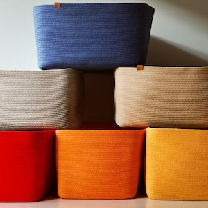 May include: A collection of rectangular storage baskets made from coiled rope, featuring a variety of solid colors including blue, beige, tan, red, orange, yellow, and green. Each basket has a small brown leather-like tab attached to the top rim.