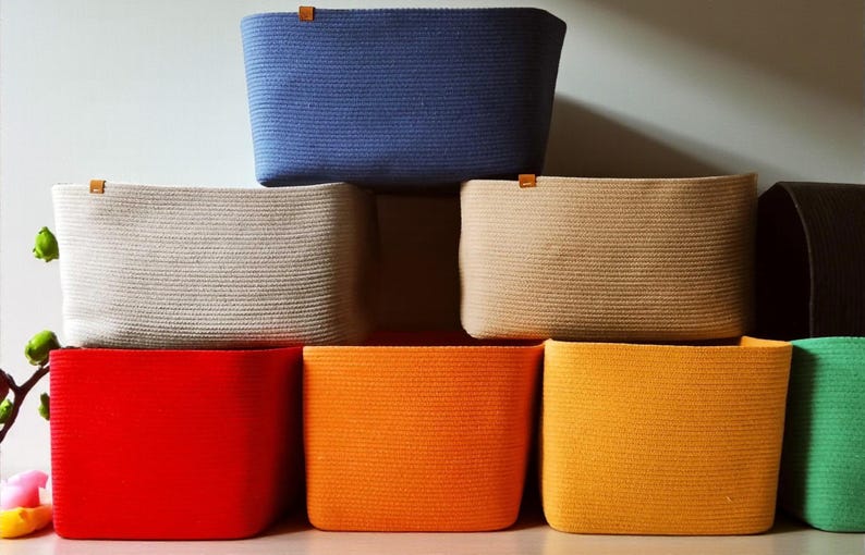 May include: A collection of rectangular storage baskets made from coiled rope, featuring a variety of solid colors including blue, beige, tan, red, orange, yellow, and green. Each basket has a small brown leather-like tab attached to the top rim.