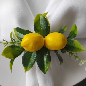 May include: A white napkin ring with two yellow lemons and green leaves. The lemons are attached to the ring with a brown twine.