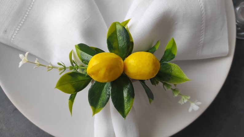 May include: A white napkin ring with two yellow lemons and green leaves. The lemons are attached to the ring with a brown twine.